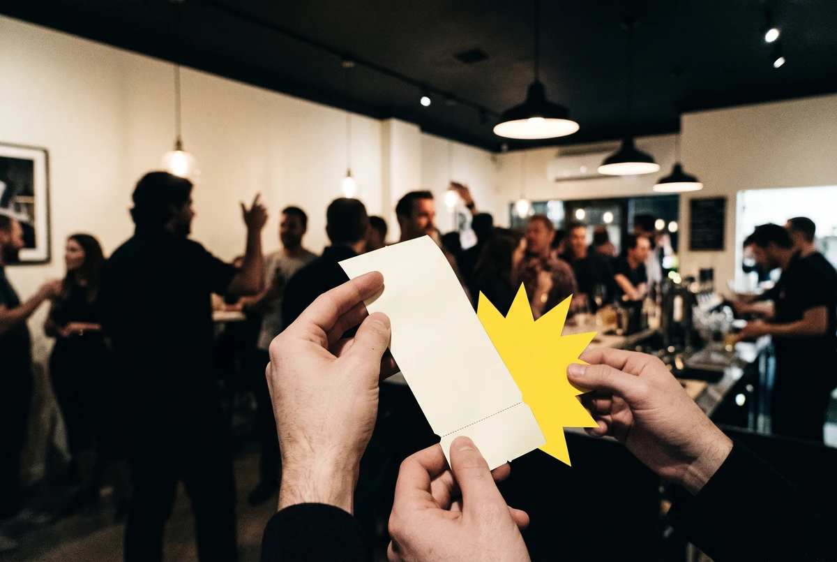 A guest's hands hold a blank receipt with a tear-off coupon along the bottom, a crowded grand-opening restaurant bar softly out of focus behind them