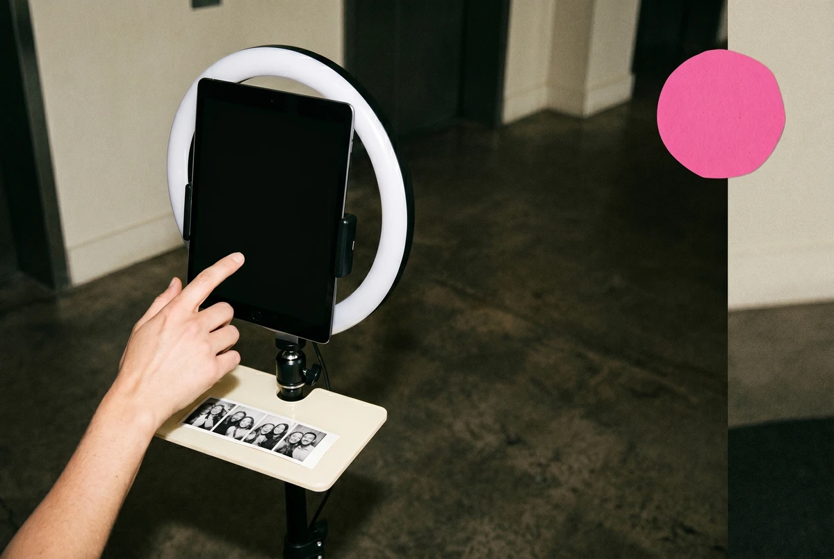 Overhead view of a guest's hand reaching toward an iPad on a ring-light stand at a hotel lobby photo station, a printed photo strip resting on a shelf below