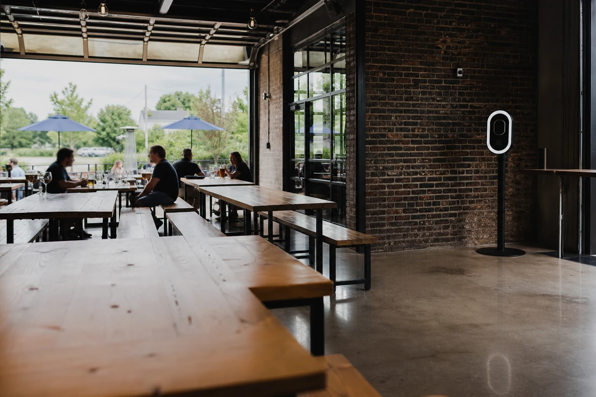 A compact iPad photo booth on a slim stand tucked into the corner of a brewery taproom, taking far less floor space than the long communal tables.