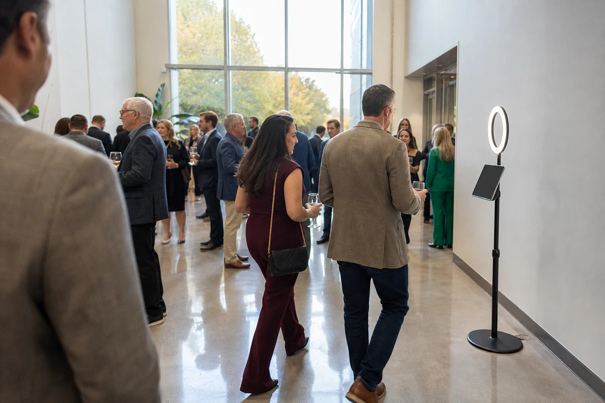An iPad photo station stands near the entrance of a bright campus atrium during an alumni reception, with open floor space and guests mingling nearby.