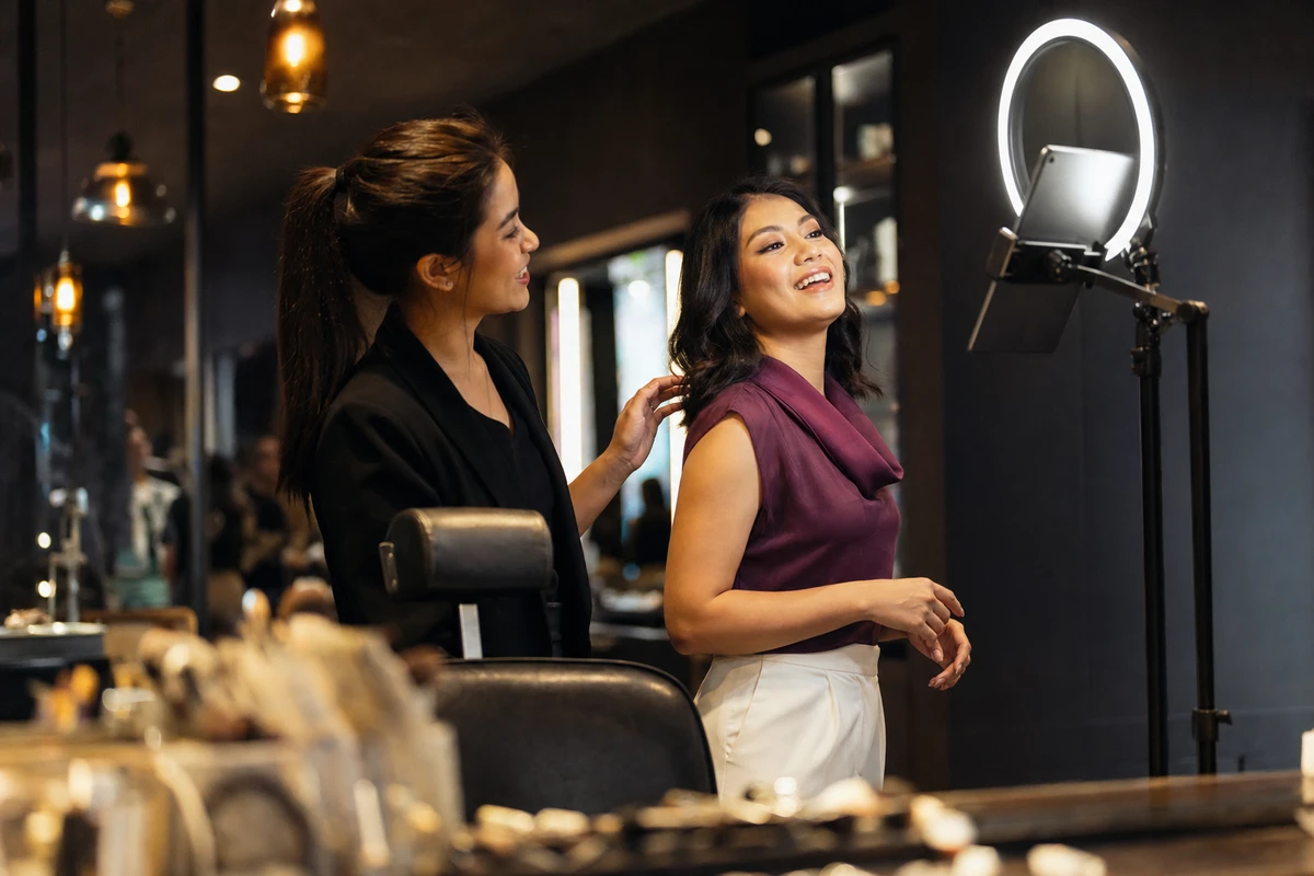 A customer poses for an iPad photo-booth capture station on a ring-light stand beside a beauty-hall makeover chair while a brand ambassador guides her pose.