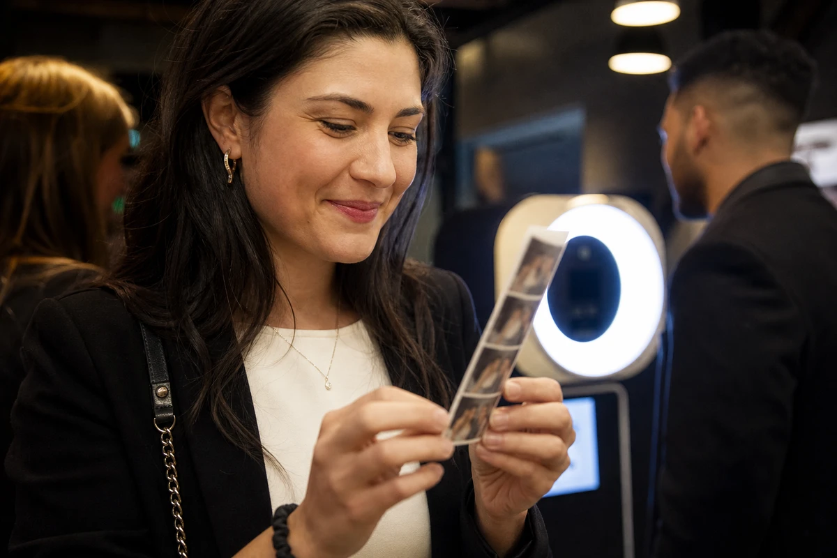 A corporate event guest holding a freshly printed photo strip and smiling as the photo booth glows out of focus behind them.