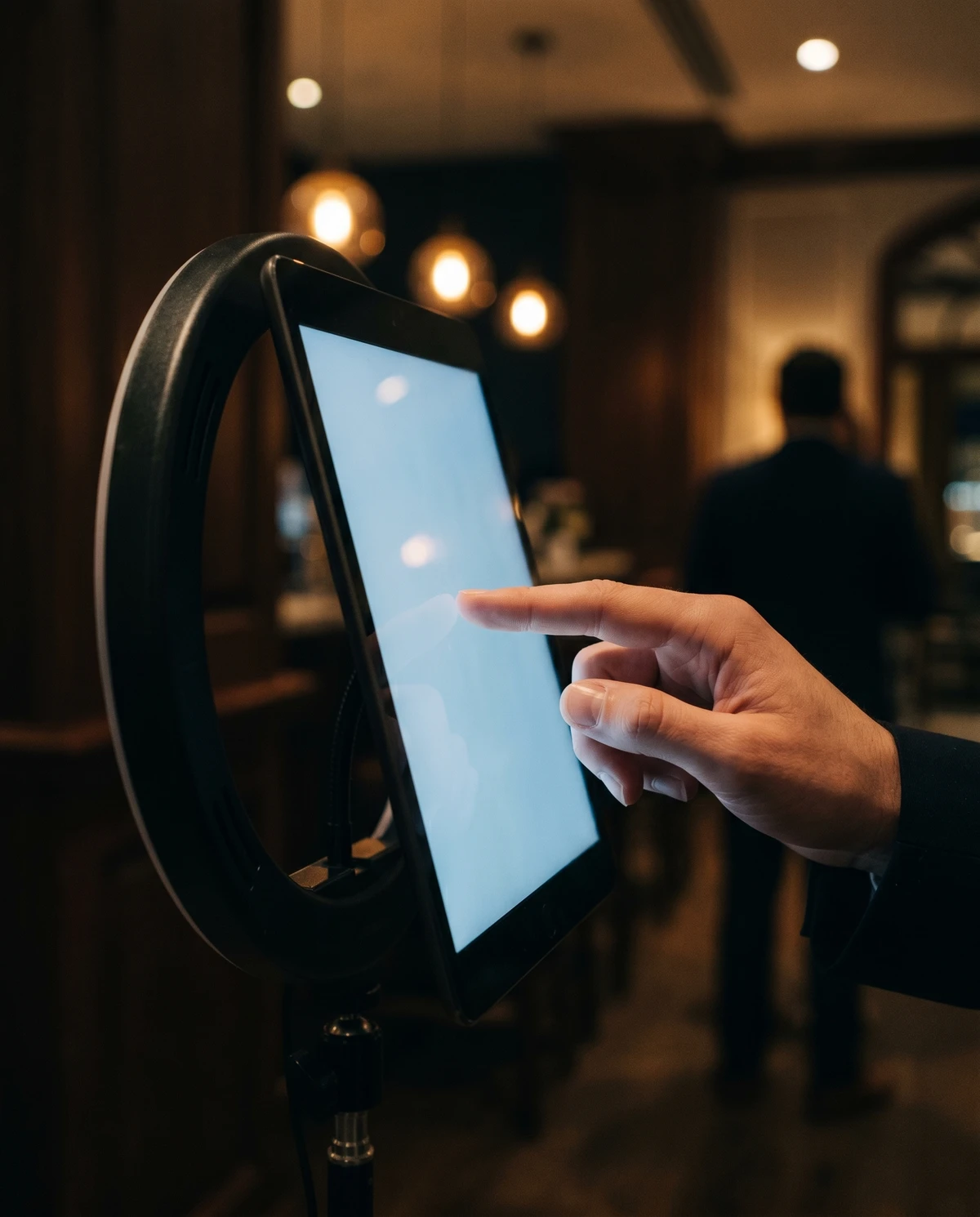 Close-up of a fingertip about to tap a glowing iPad screen mounted on a ring-light stand inside a dim hotel-bar venue, warm pendant lights blurred behind.