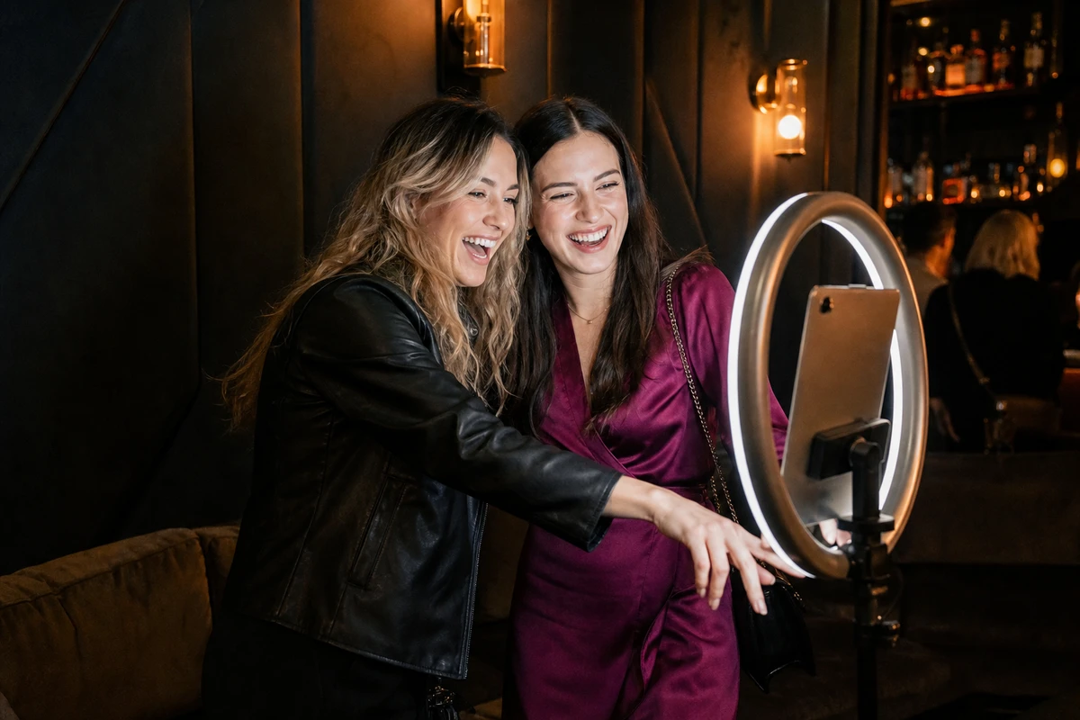 Two friends leaning in together and laughing as they pose at a photo-booth capture station in the lounge corner of a bar.