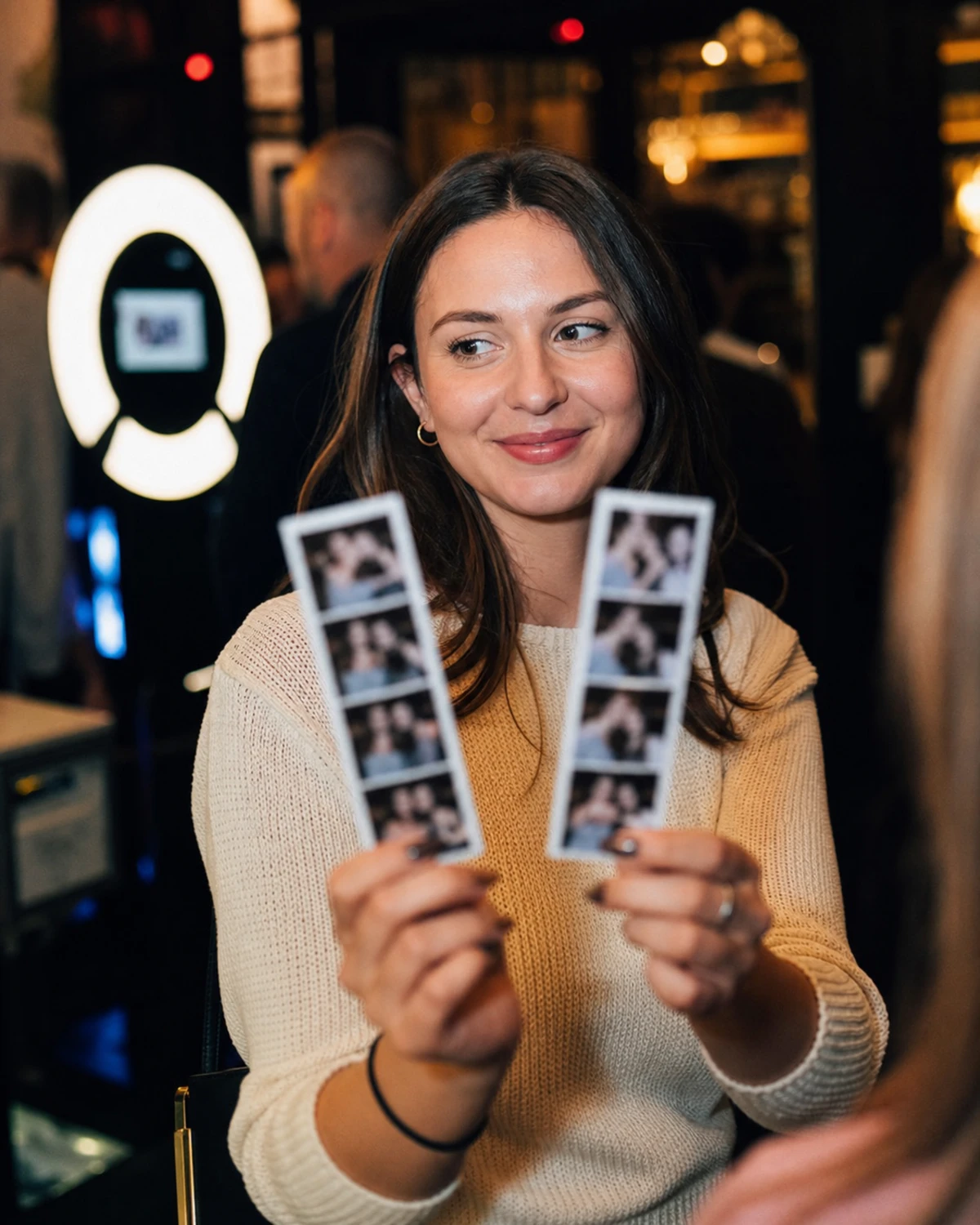 A guest holds two freshly printed photo strips at chest height after a photo-booth session, turning to show a friend off-frame.