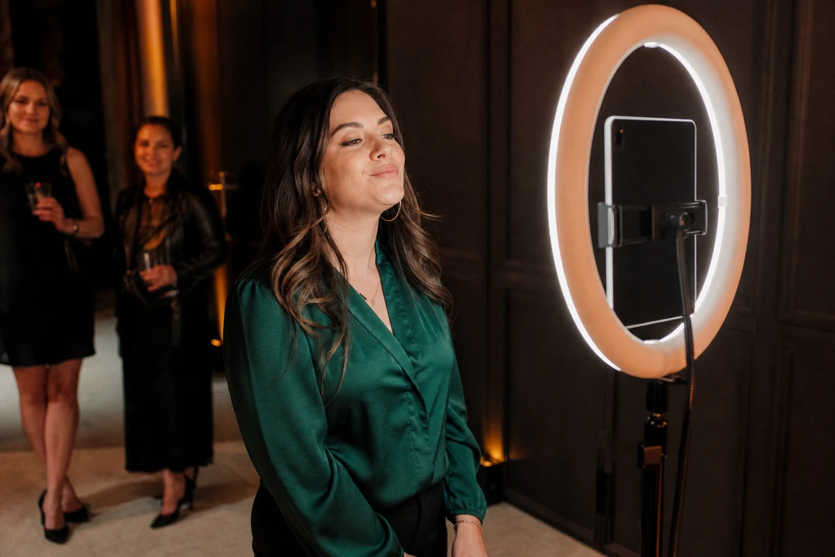 A woman poses for a photo at an iPad photo booth on a ring-light stand during an evening event activation, with two guests waiting in the background.