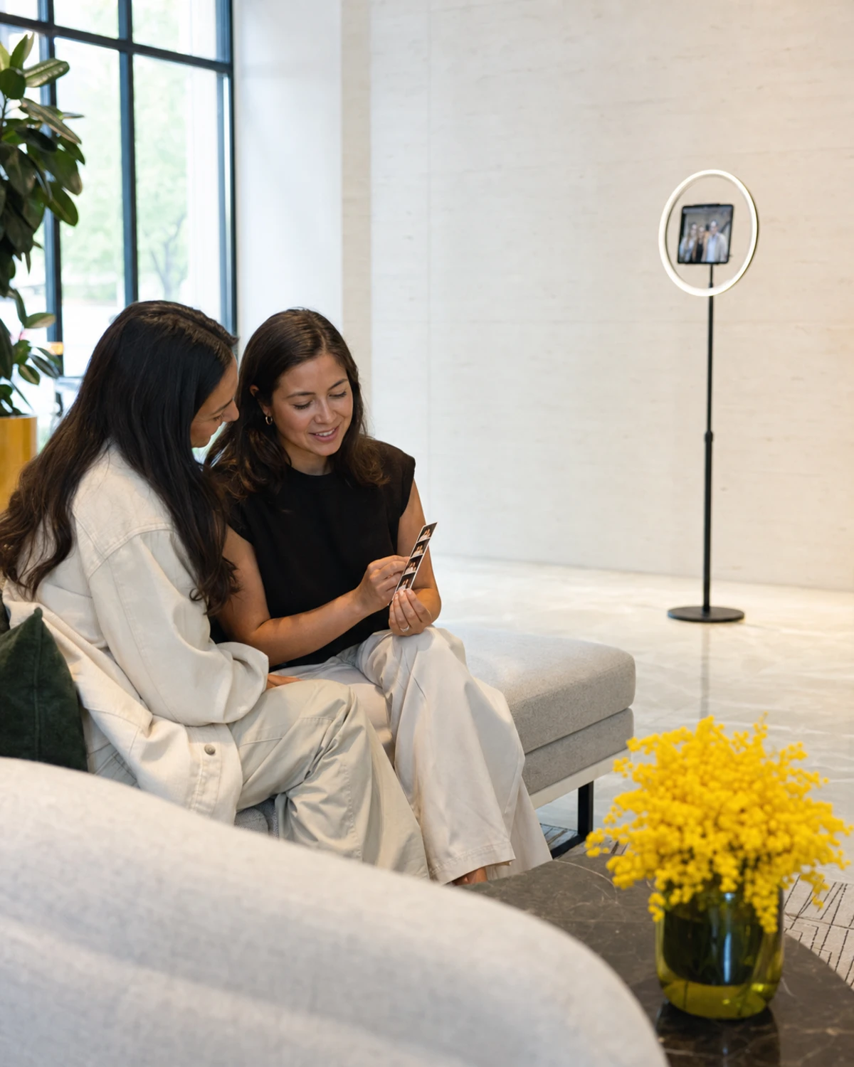 Two hotel guests on a lobby sofa share a freshly printed photo strip while a photo-booth station stands nearby.