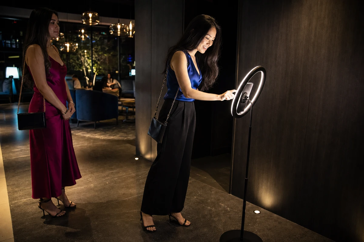 A single hotel guest leaning in to tap the screen of a branded photo station mid photo session in a hotel lounge corner