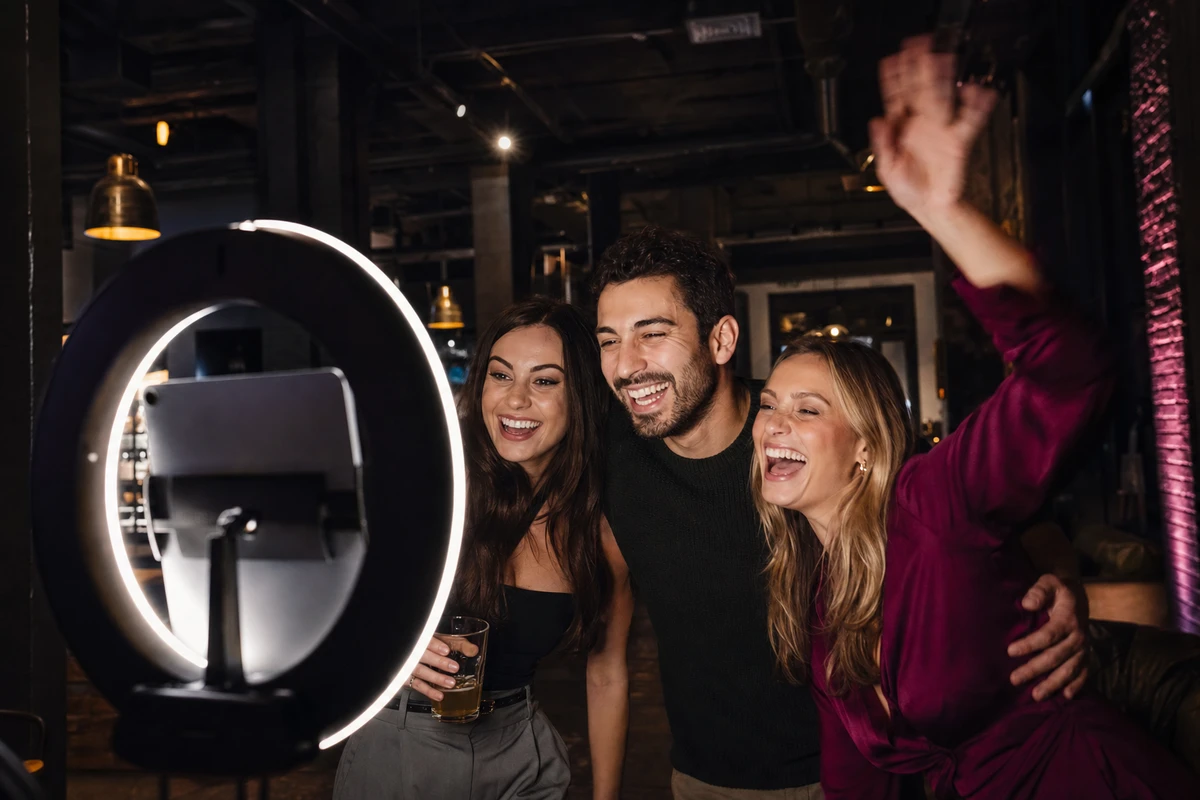 Three friends laughing together at an unattended iPad photo booth in a dimly lit brewpub during an evening event.