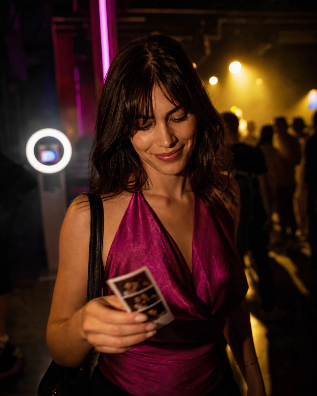 A nightclub guest walking back toward the dancefloor holding a freshly printed photo strip and glancing down at it with a small smile.