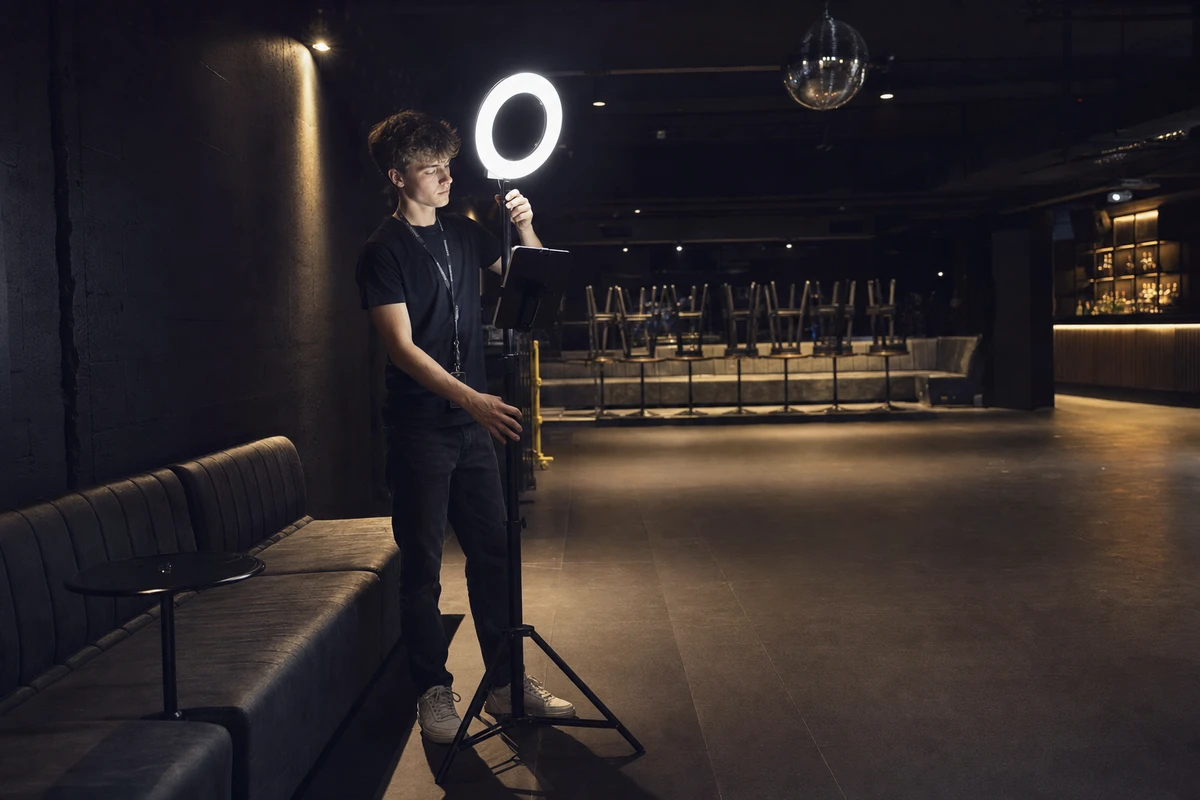 A photo-booth operator adjusting the ring light of an iPad photo station inside a nightclub before the doors open, house lights partly up and the dancefloor empty.