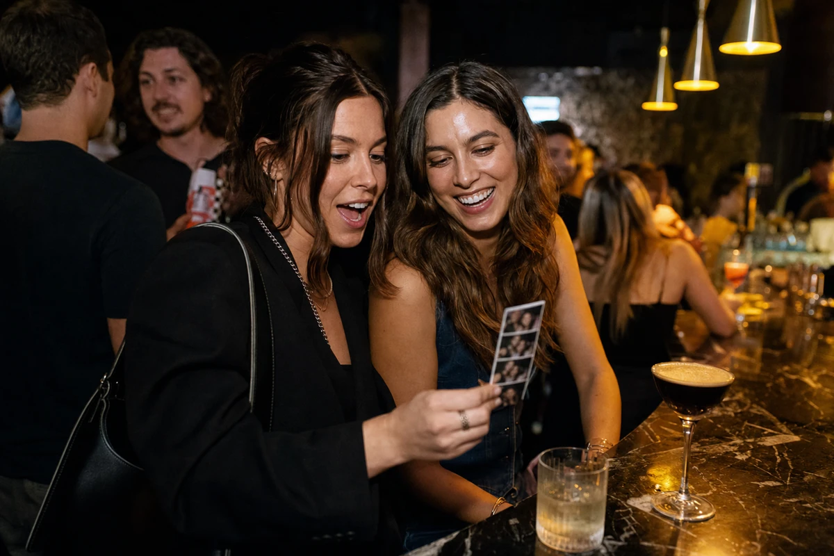 A bar guest holding up a freshly printed photo strip to show a smiling friend, both looking down at it under warm pendant light at the end of the bar.