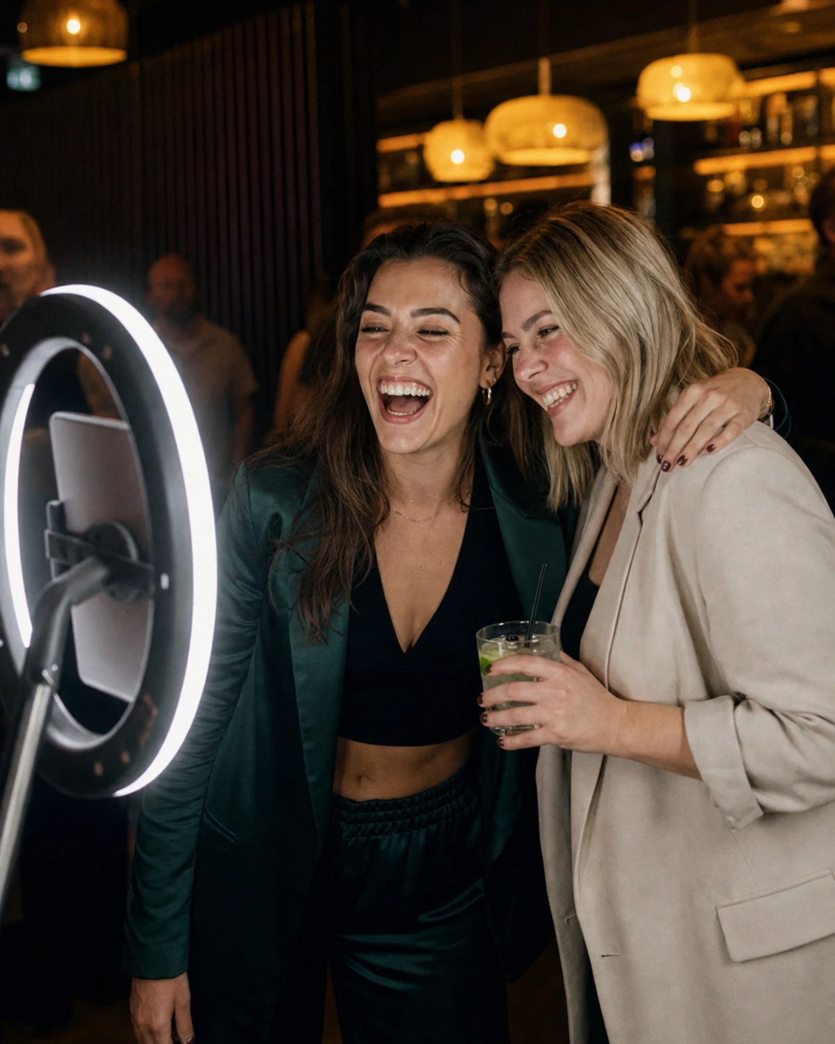 Two friends standing together and laughing at a photo-booth station in a bar lounge, the ring light glowing on their faces as the photo is taken.