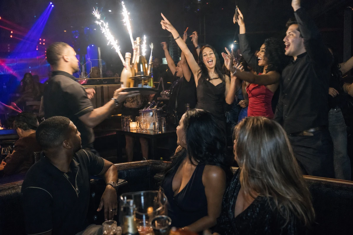 A server carries sparkler-topped bottles to a standing, cheering VIP group while guests at the next banquette turn to watch the reveal.