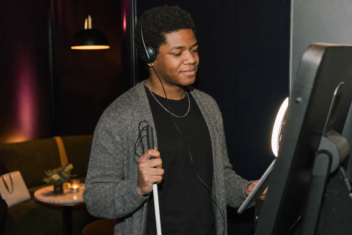 A guest wearing headphones for private audio holds a pose at a photo booth, using it independently with a folded white cane at their side.