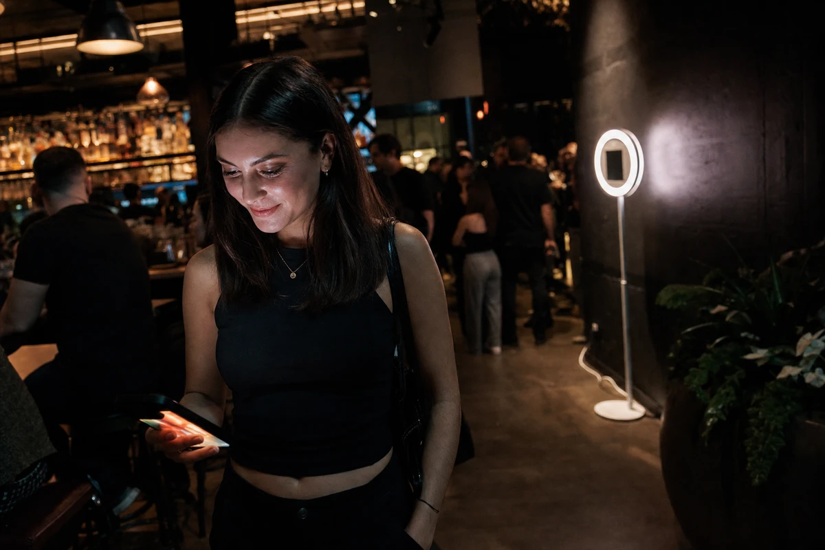 A bar guest standing a step away from a tablet photo booth at night, smiling at her phone after receiving the digital copy of her photo.