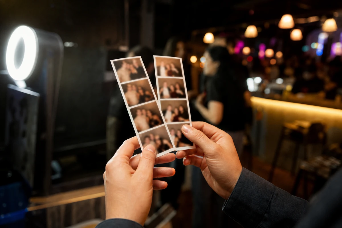 A guest's hands holding two freshly printed photo strips just collected from a bar's photo booth, the bar glowing softly out of focus behind.