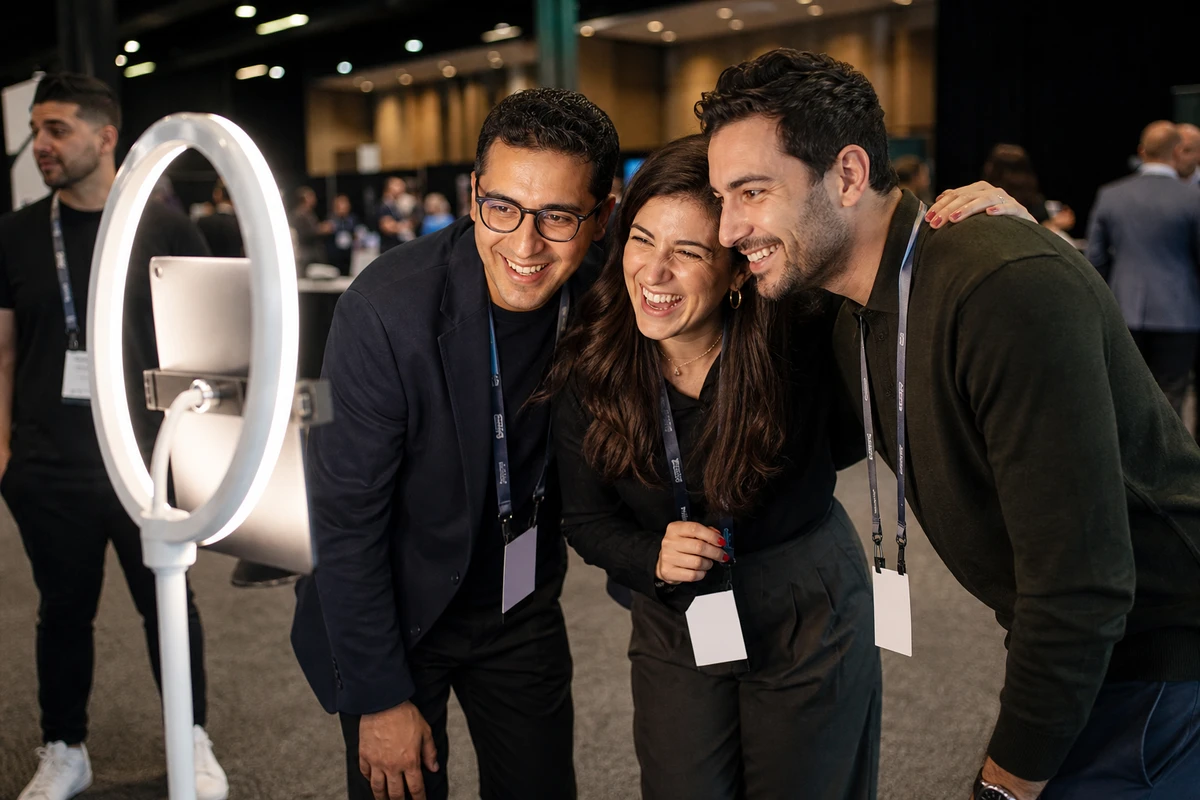 Three conference professionals in lanyards laugh and pose together at a photo booth on a ring-light stand during a trade-show activation.