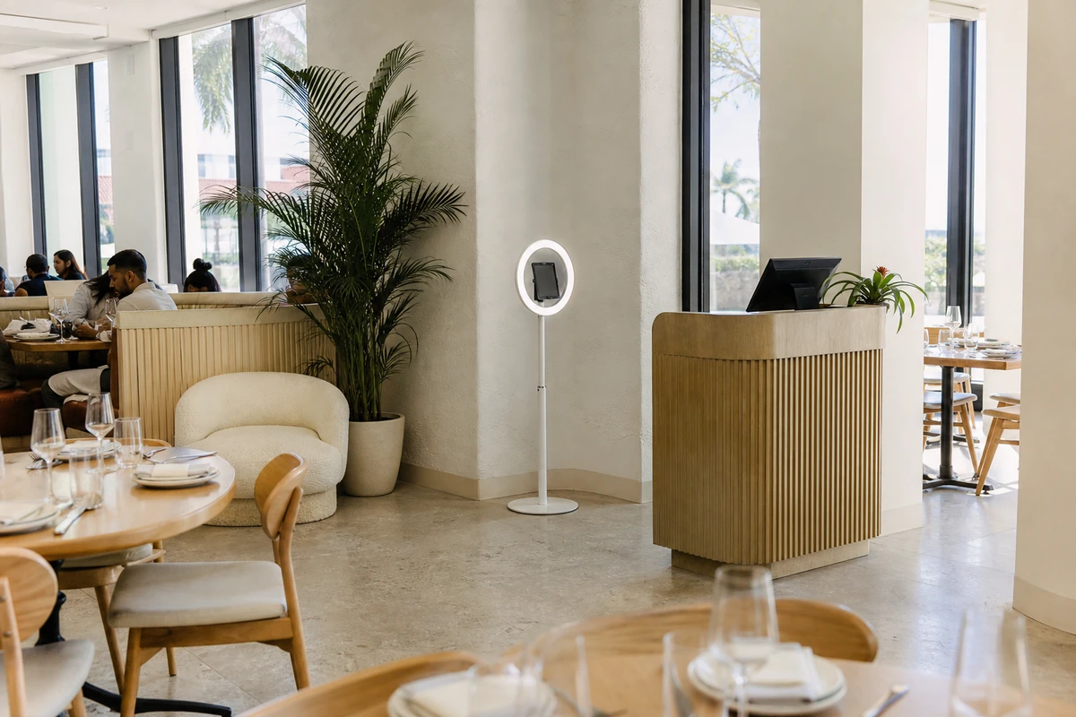 A photo booth on a ring-light stand installed as a permanent fixture in the lounge corner of a daylit restaurant near the host stand, with clear floor space around it.