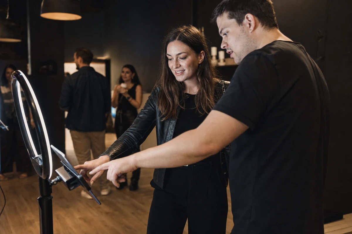 A guest taps a photo booth tablet to opt in while an operator points and explains at a brand activation