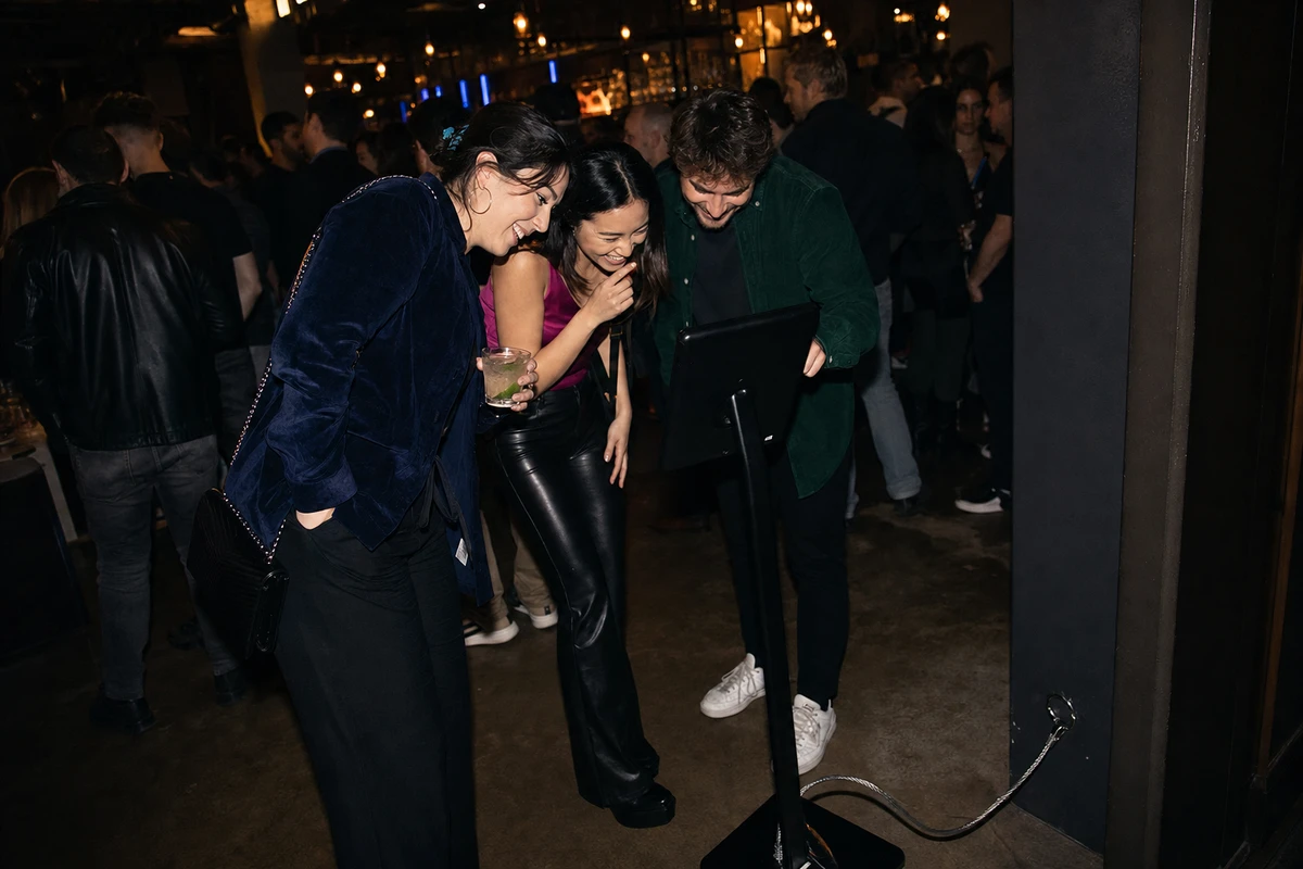 Three friends gather at a tethered iPad photo-booth kiosk in a crowded dark bar at night, the steel security cable visible anchoring its base.