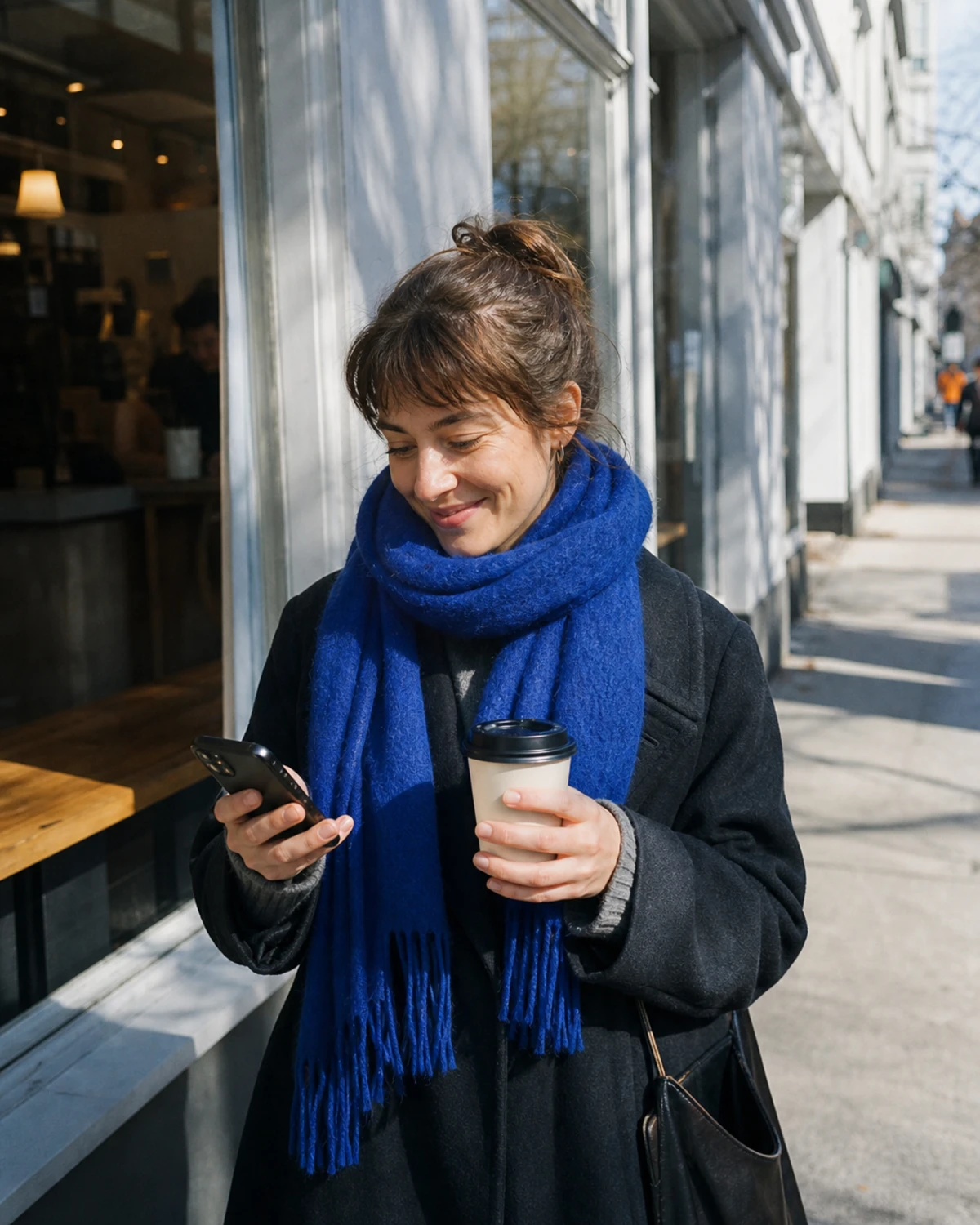 A coffee shop regular stands on the sidewalk outside, smiling privately at her phone after receiving her photo from the booth.