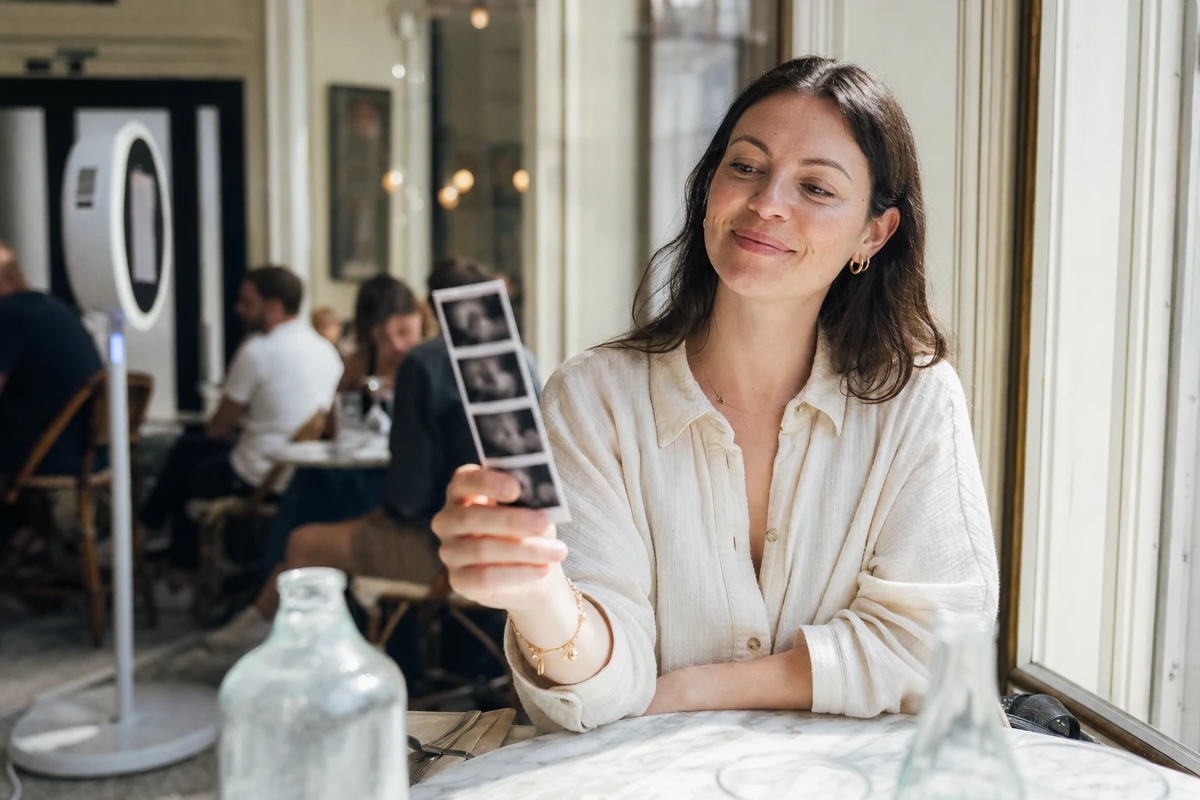 A restaurant guest sits by a sunlit window and smiles at a freshly printed photo strip held in her hand.