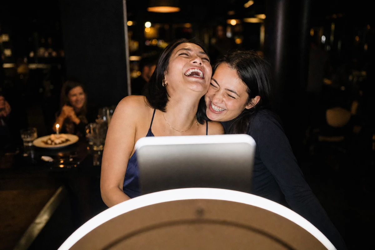 Two restaurant guests laugh cheek to cheek inside a photo booth's ring light, a dessert with a single lit candle glowing on the table behind them.