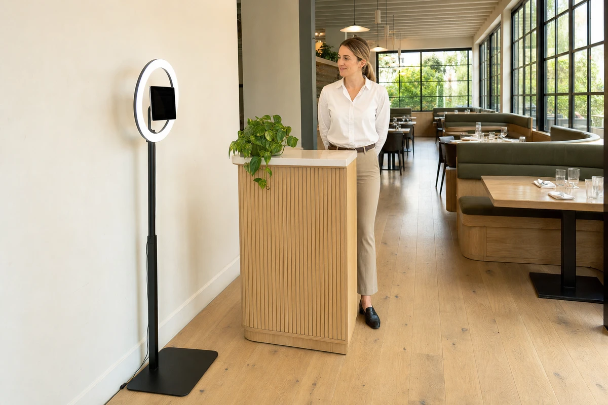 A tablet photo station stands on open floor beside the host stand of a daylit restaurant before service, showing where a venue places its capture surface.