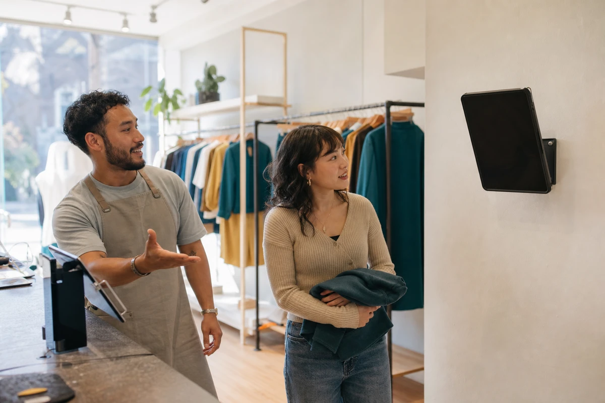 A store associate near the register gestures toward a wall-mounted selfie station, inviting a customer holding a garment to use it before leaving.