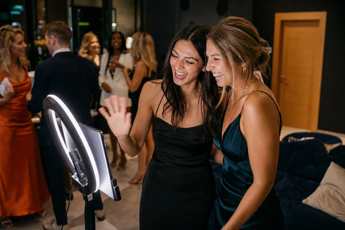 Two med spa patients laughing together at a self-serve iPad photo station lit by its ring light during an evening VIP client night.